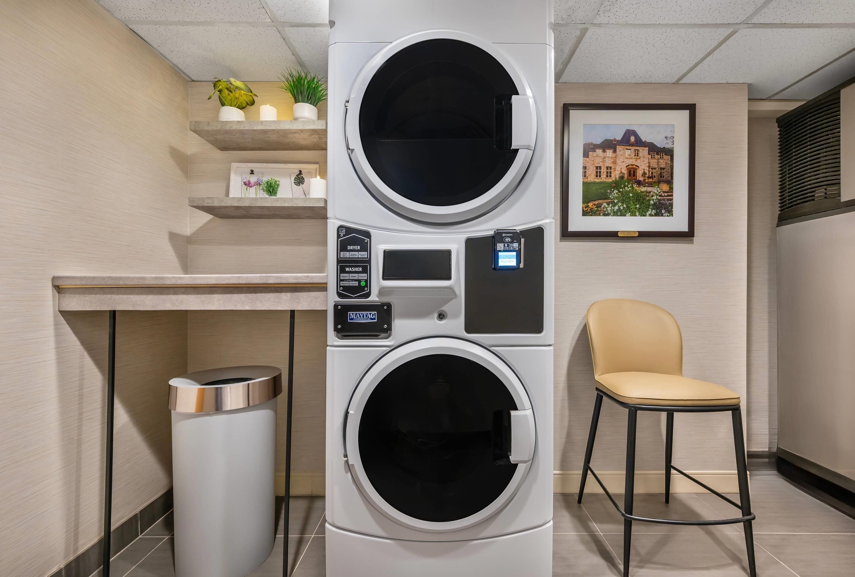 A modern laundry room with a large wall-mounted washer and dryer, a small trash can, a chair, and a framed picture on the wall.