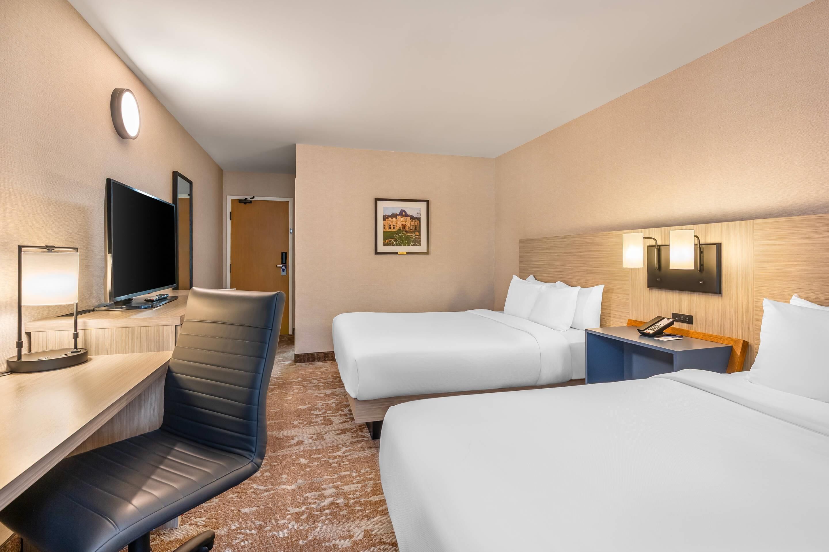 Une chambre d’hôtel avec deux lits, un bureau avec une chaise, une télévision et un tableau encadré au mur.