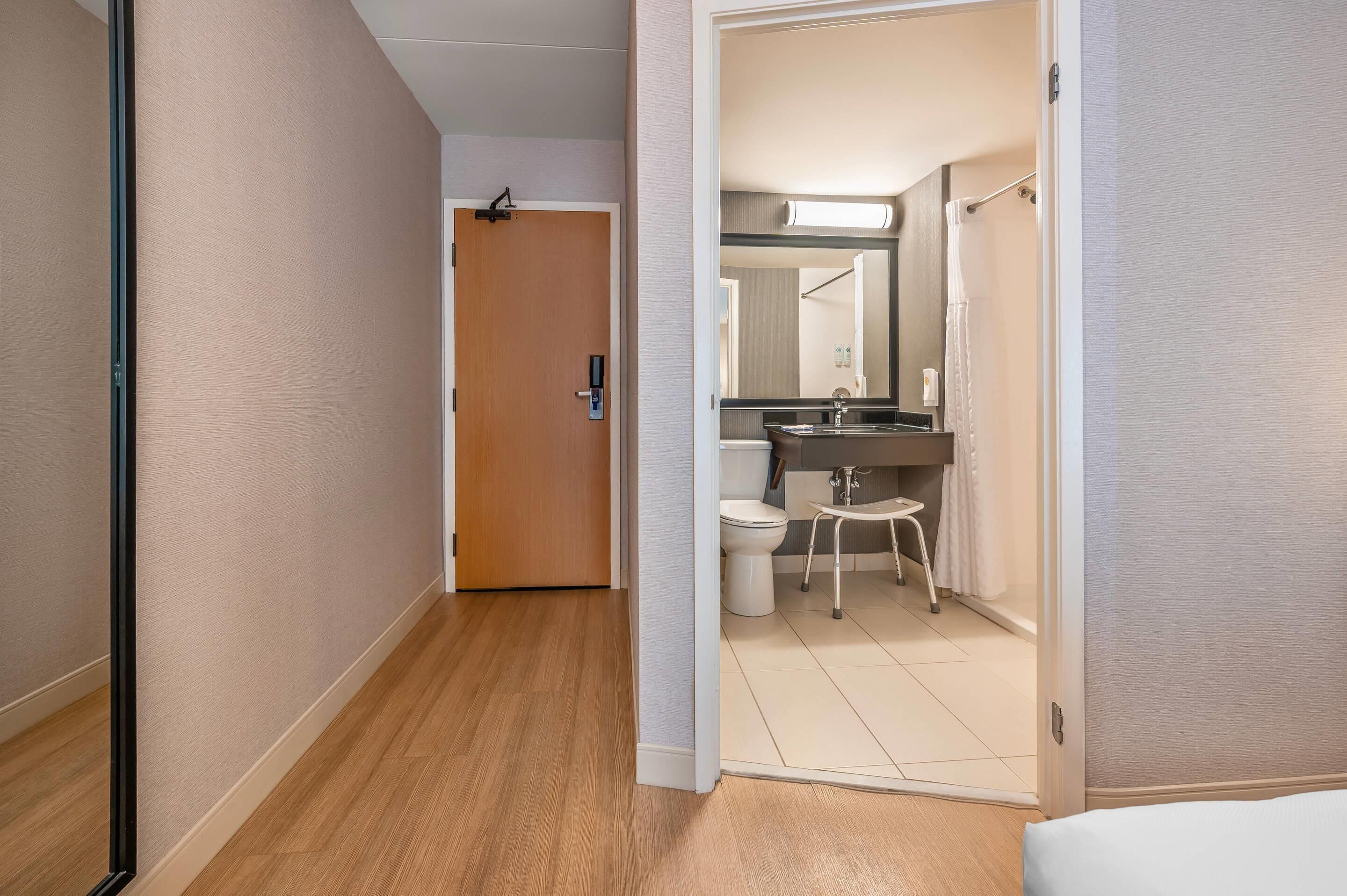 A hotel hallway with a wooden floor, a door leading to a bathroom with a sink, toilet, and mirror.