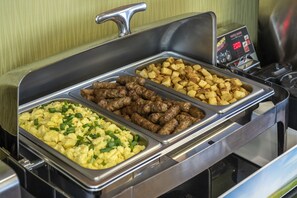 A buffet with three trays of food: scrambled eggs, sausages, and roasted potatoes.