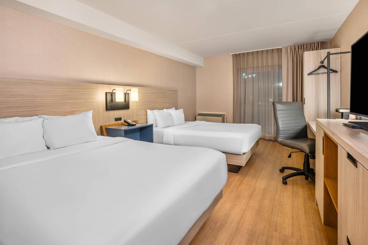 A hotel room with two beds, a desk with a chair, a TV, and a wall-mounted lamp.