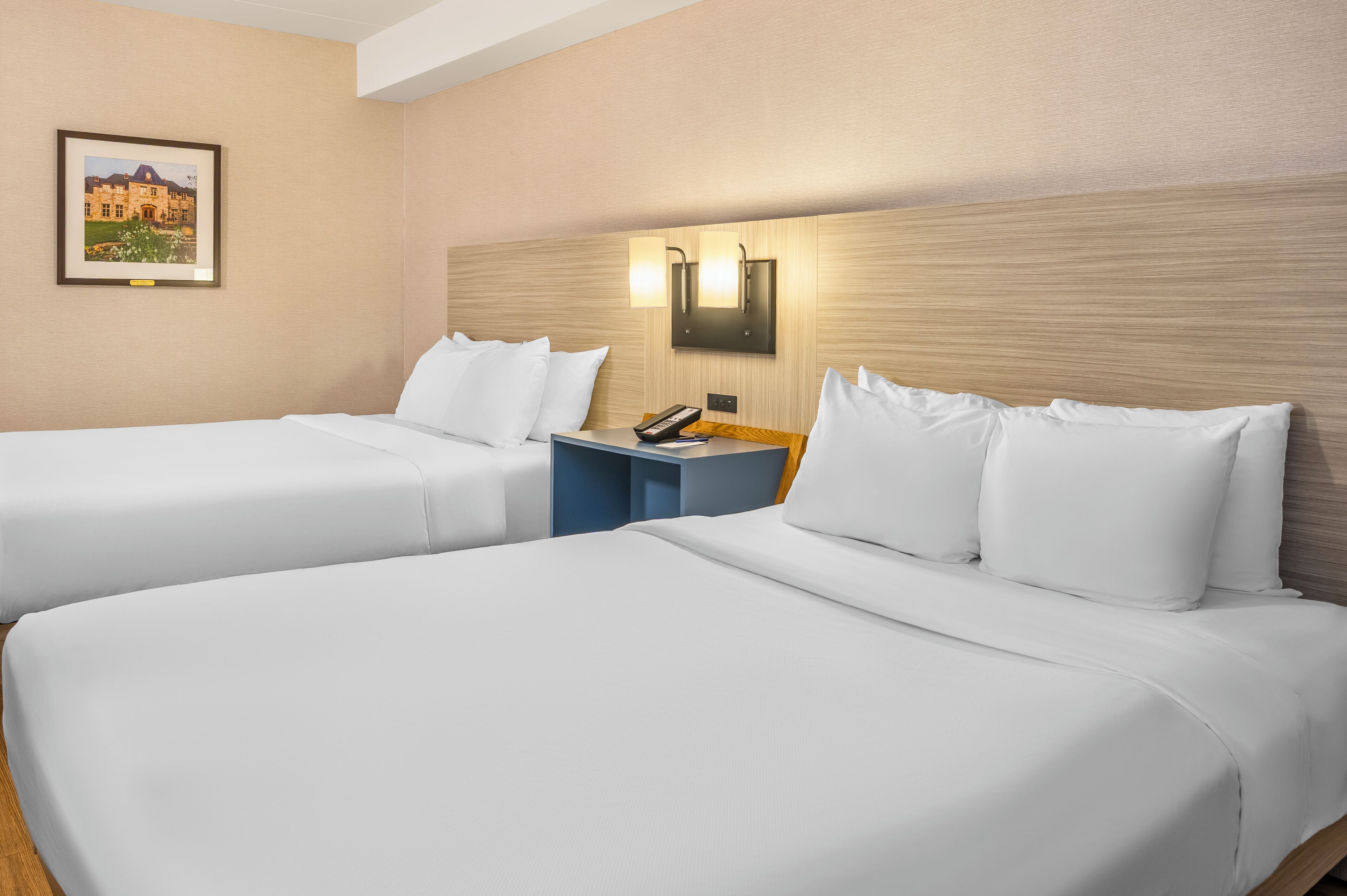 A hotel room with two beds, a nightstand with a phone, and a wall-mounted light fixture.