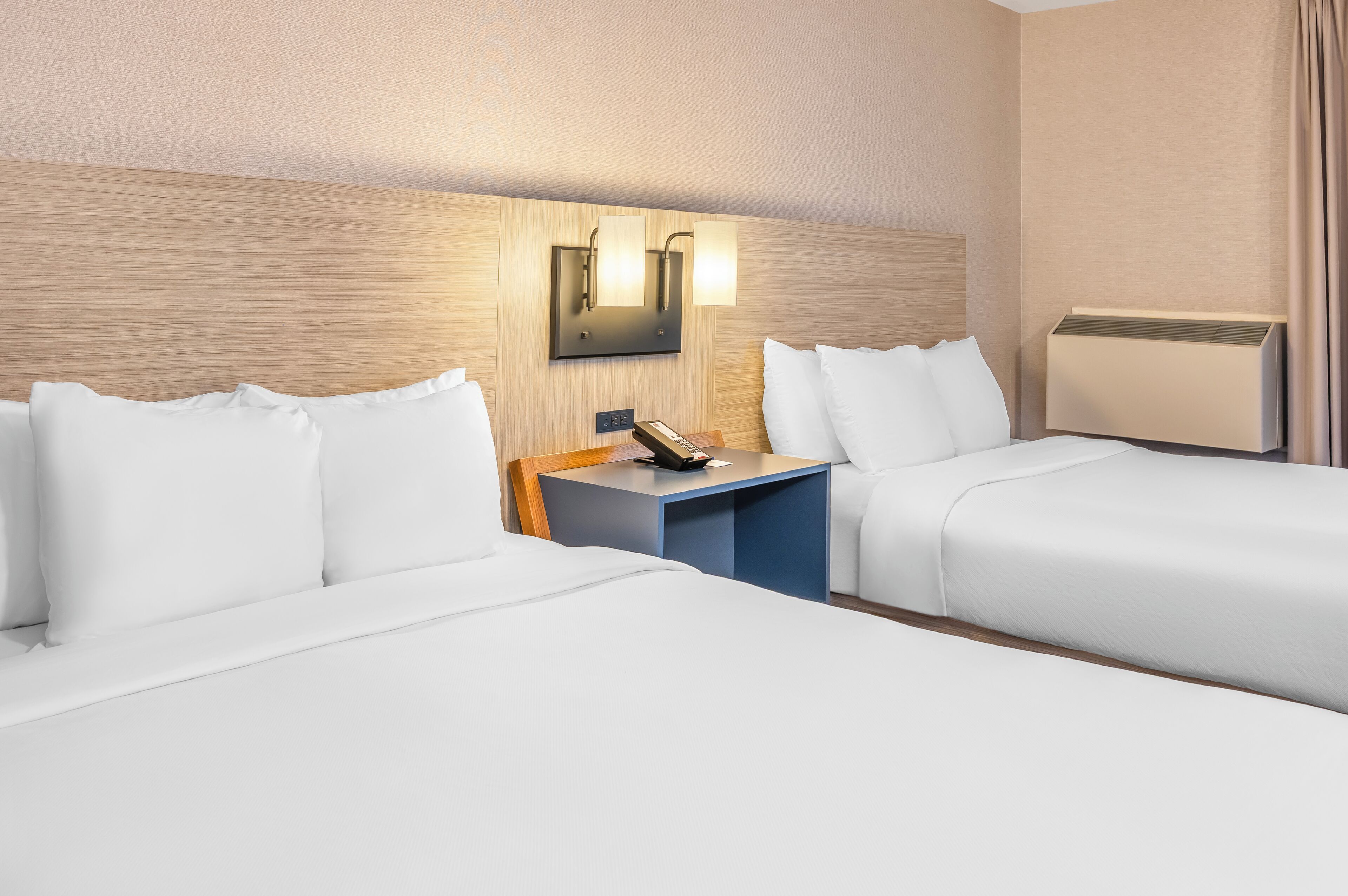 A hotel room with two beds, a nightstand with a phone, and a wall-mounted light fixture.