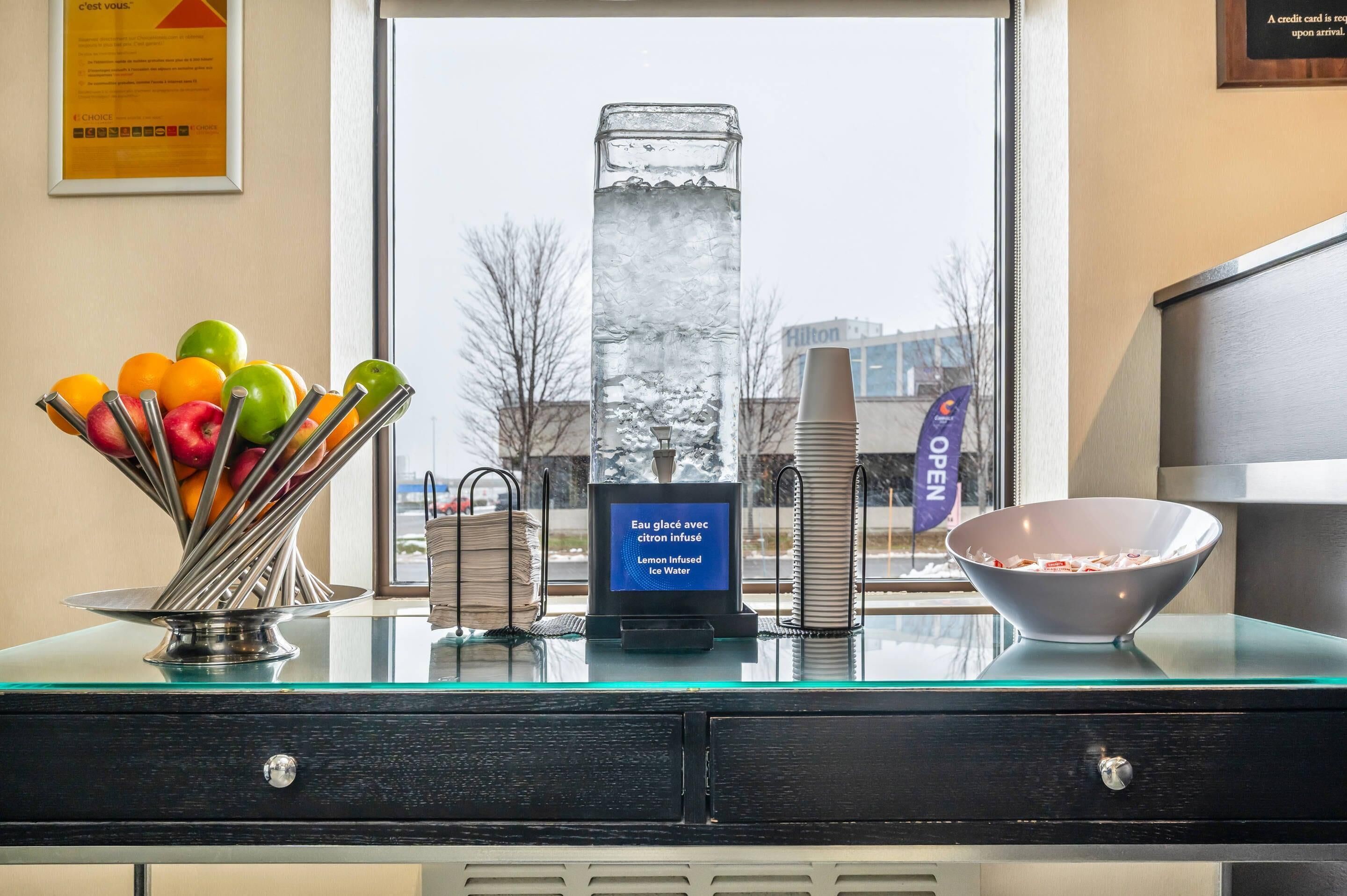 A hotel reception desk with a glass display case containing a fruit bowl, a lemon-infused ice water dispenser, and a bowl of cereal.