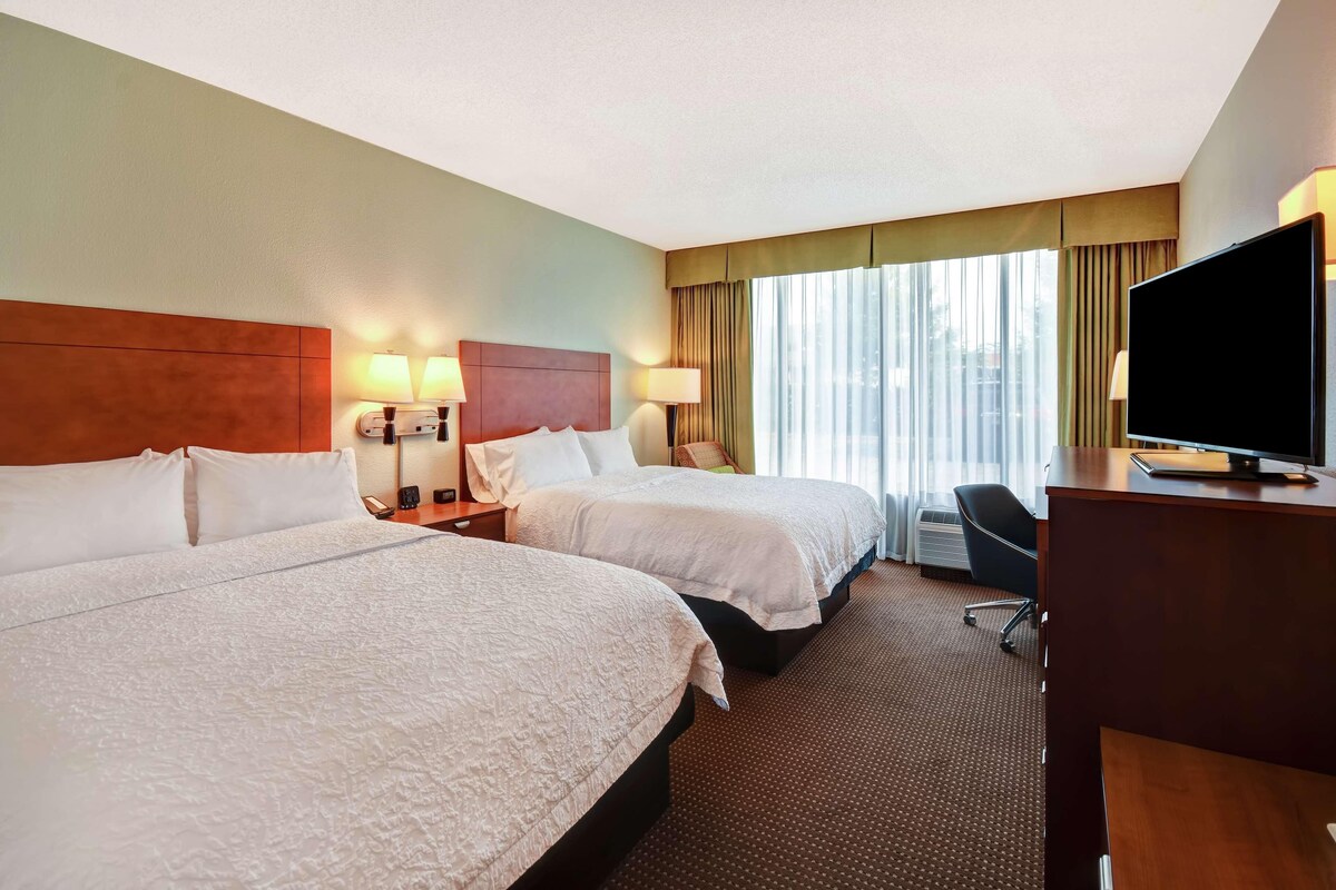 A hotel room with two beds, a desk with a TV, and a window with curtains.
