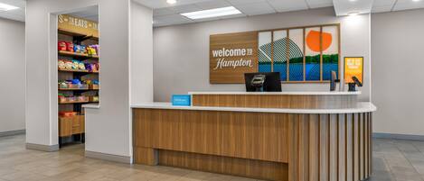 A hotel reception desk with a 'Welcome to Hampton' sign, a self-service kiosk, and a snack shelf.