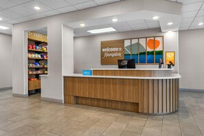 A hotel reception desk with a 'Welcome to Hampton' sign, a self-service kiosk, and a snack shelf.