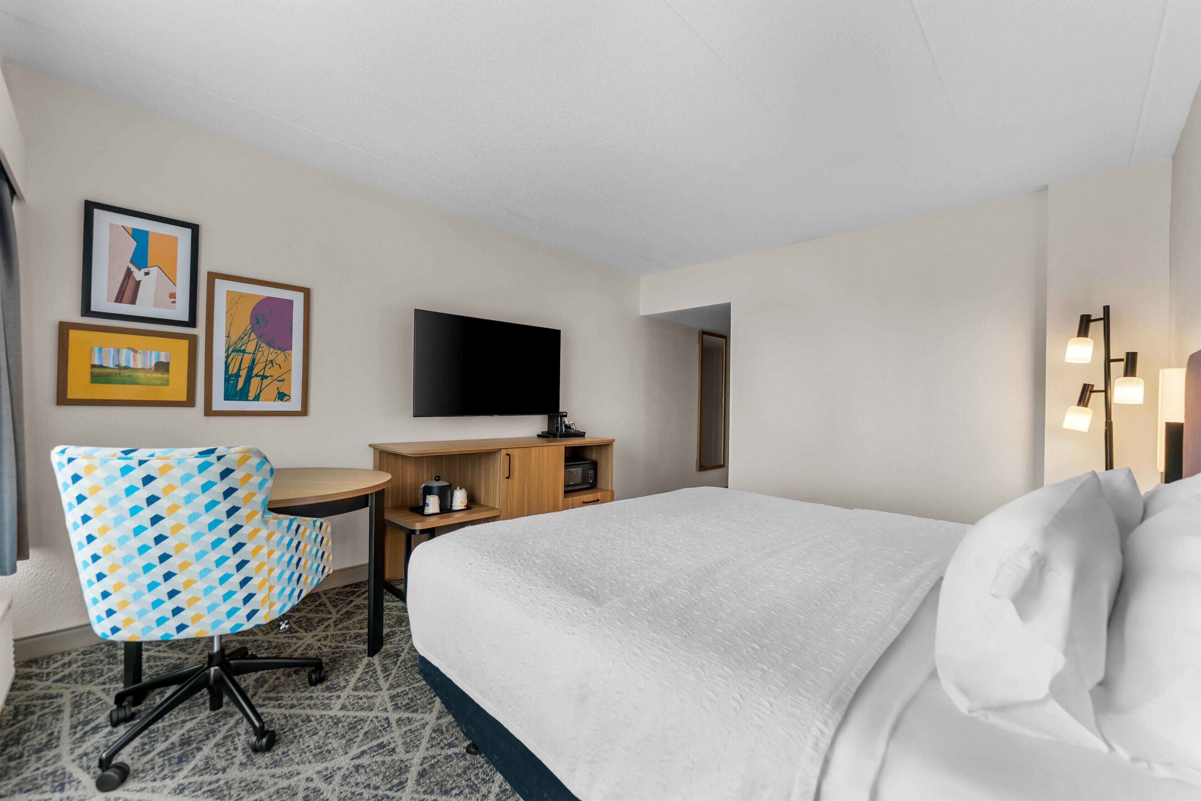 A hotel room with a bed, a desk, a chair, a TV, and artwork on the wall.