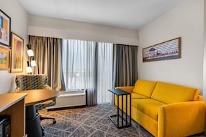 Une chambre d’hôtel avec un bureau, un canapé jaune, une moquette à motifs, une fenêtre avec des rideaux et un tableau encadré au mur.