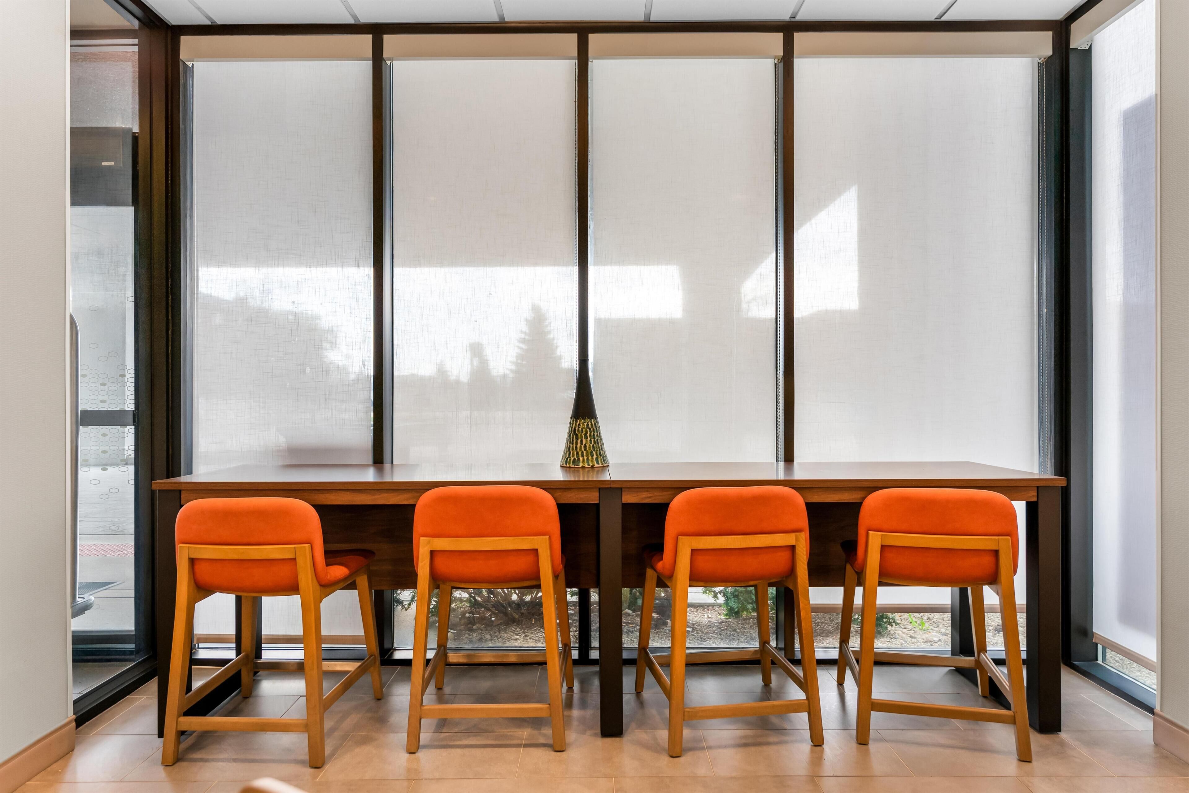 A dining area with a wooden table and four orange chairs, large windows, and a vase on the table.