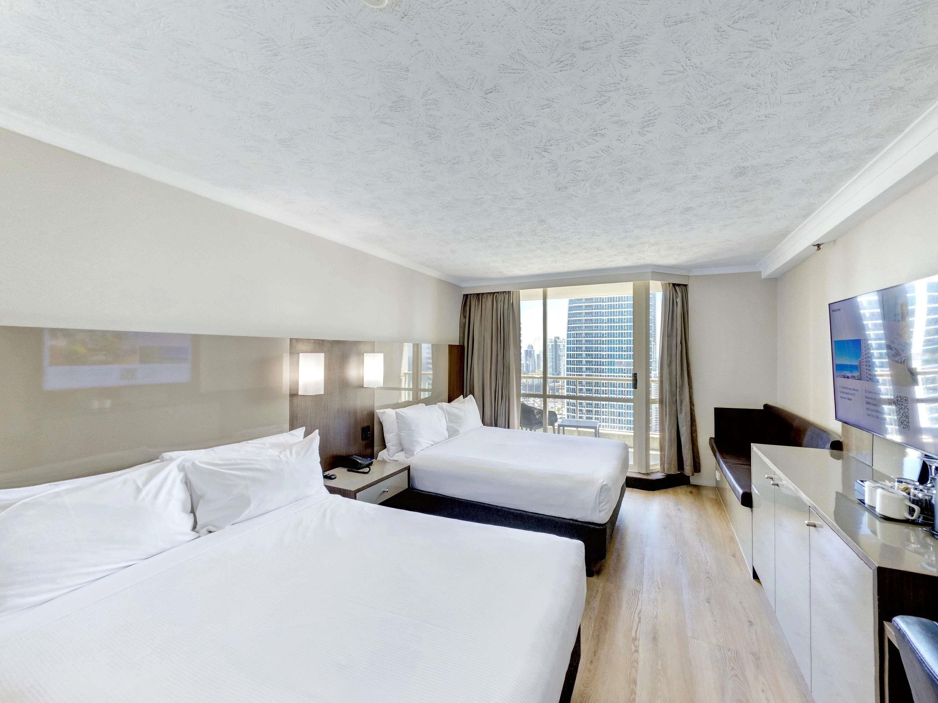 Habitación de hotel con dos camas, un escritorio, un televisor y un ventanal con vista a la ciudad.