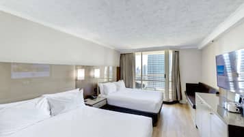 A hotel room with two beds, a desk, a TV, and a large window with a city view.