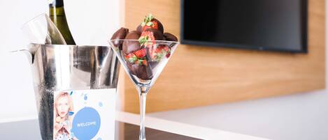 A martini glass with chocolate-covered strawberries on a white table, a bottle of champagne in a silver bucket, and a welcome card with a photo.