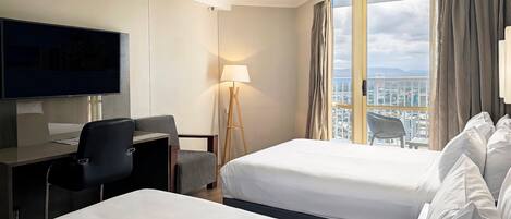 Habitación de hotel con dos camas, un escritorio con silla, televisión, lámpara de pie y vistas a la ciudad a través de amplios ventanales.