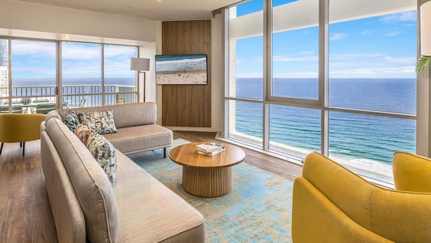A modern living room with a large window overlooking the ocean, a sofa, a coffee table, and a TV mounted on the wall. - Novotel Surfers Paradise (Surfers Paradise)