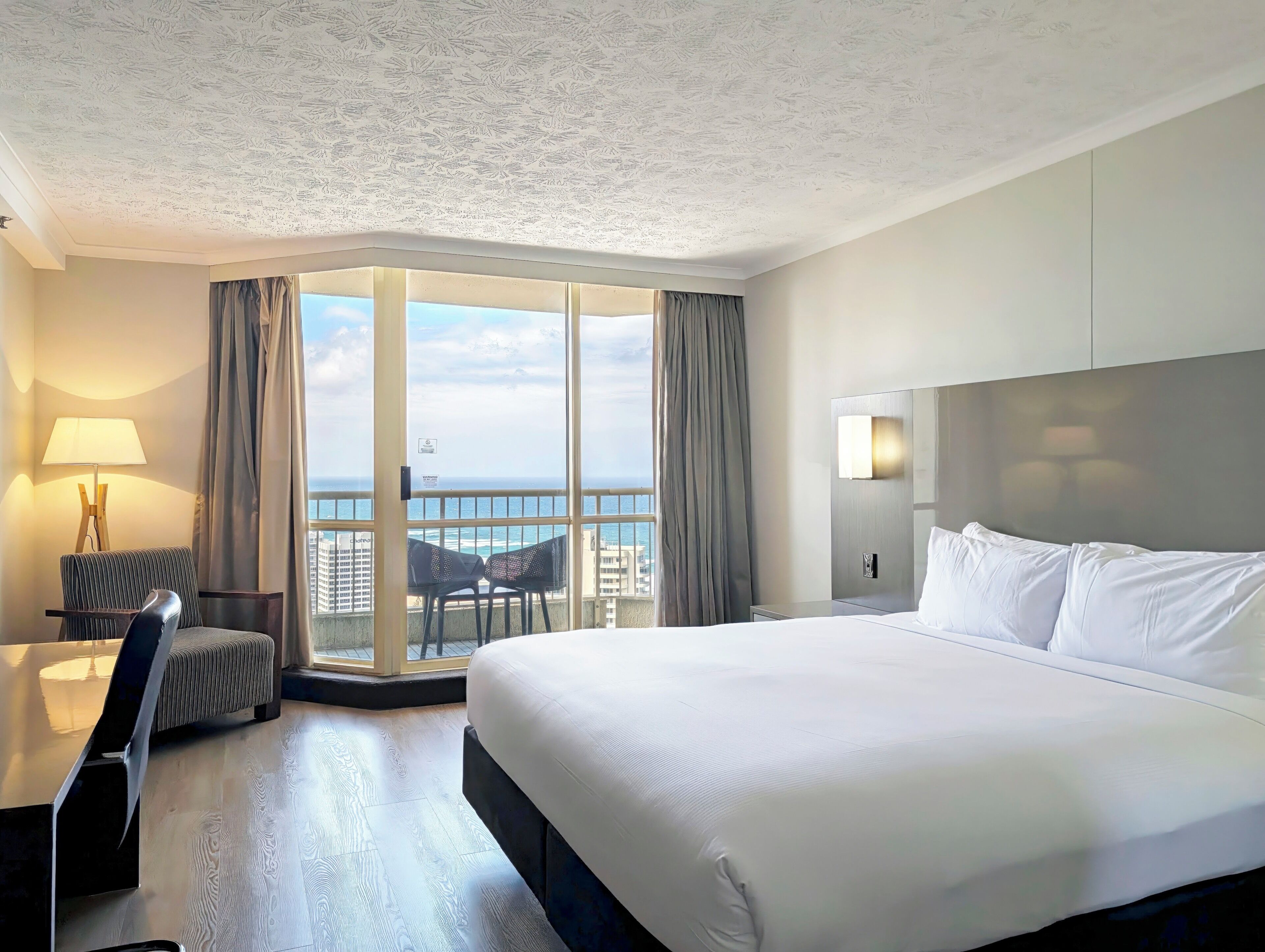 A hotel room with a large bed, a desk with a lamp, a chair, and a view of the ocean.