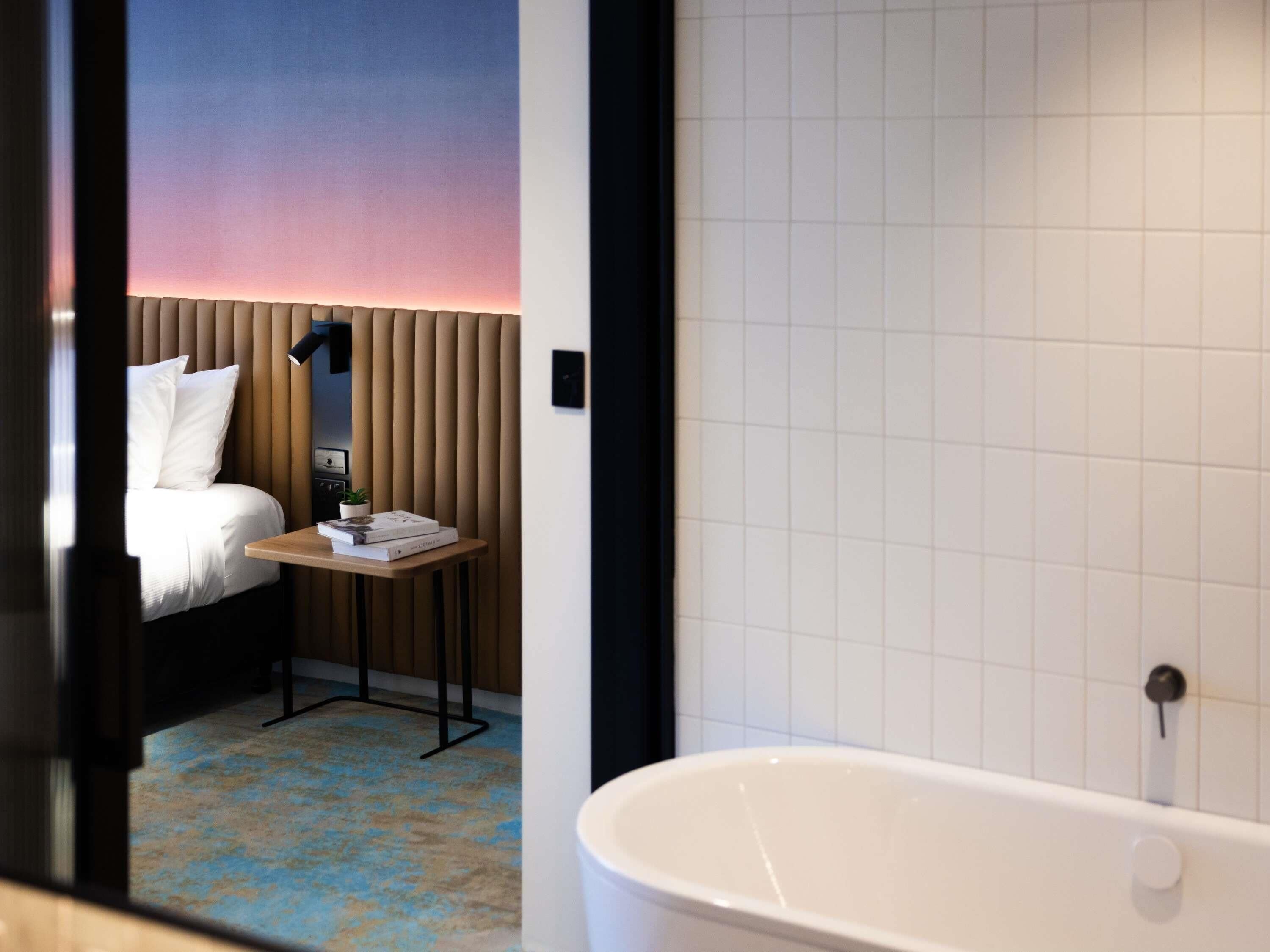A hotel room with a bed, a small table with a lamp, a potted plant, and a bathtub.