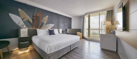 A modern hotel room with a large bed, a nightstand, a bedside table, a lamp, a mirror, and a view of the city through large windows.