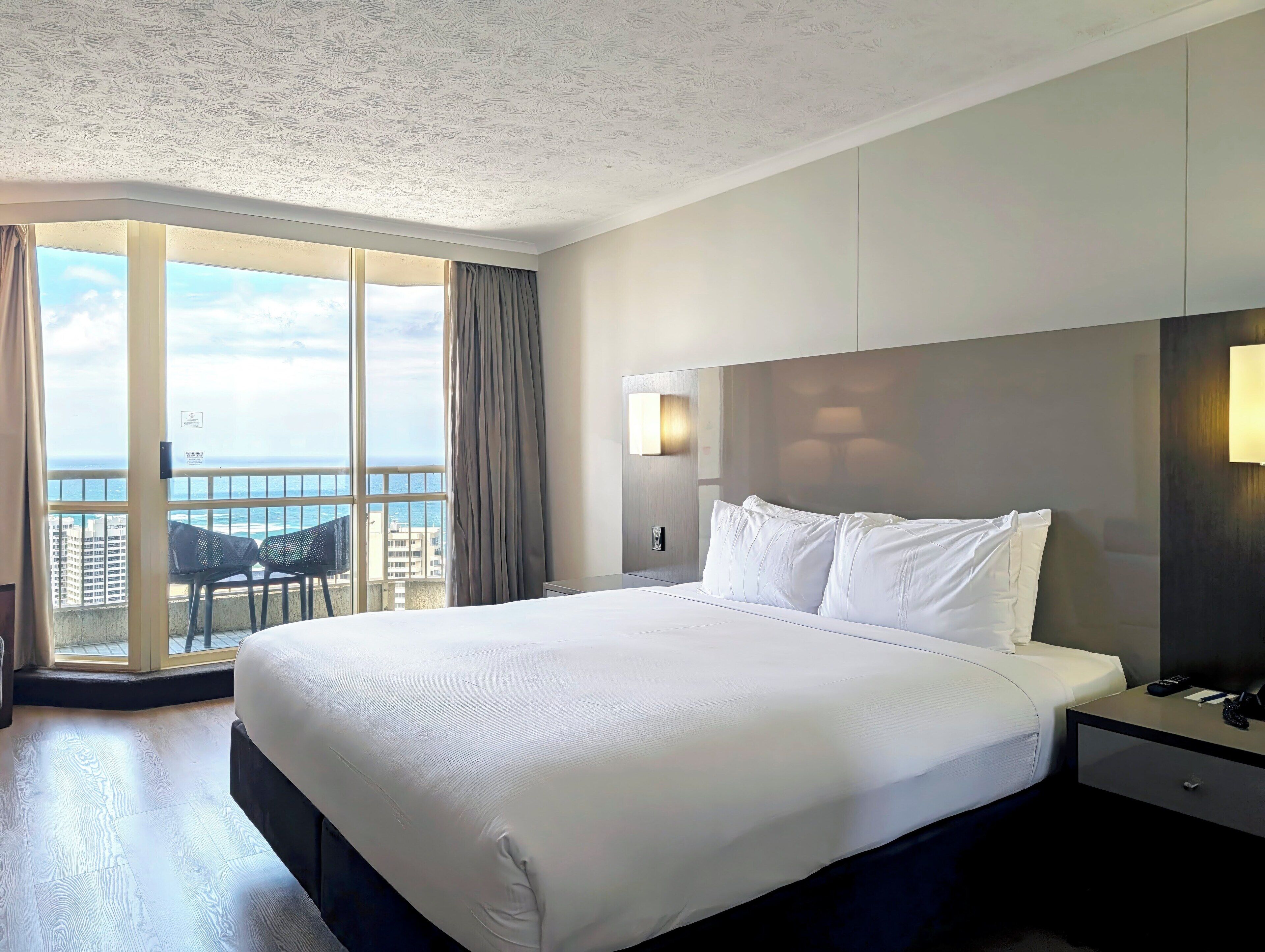 Quarto de hotel com uma cama grande, uma mesa de cabeceira, uma luminária e vista para o oceano através de uma janela ampla.