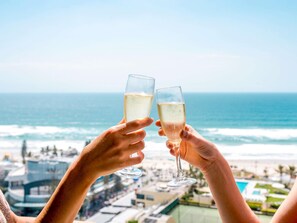 Two hands holding champagne glasses with a beach and ocean view in the background.