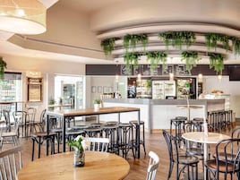 Una cafetería moderna con mesas redondas y sillas negras, una barra central y plantas colgantes.