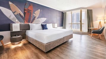 A modern hotel room with a large bed, a nightstand, a lamp, and a view of the city through large windows.