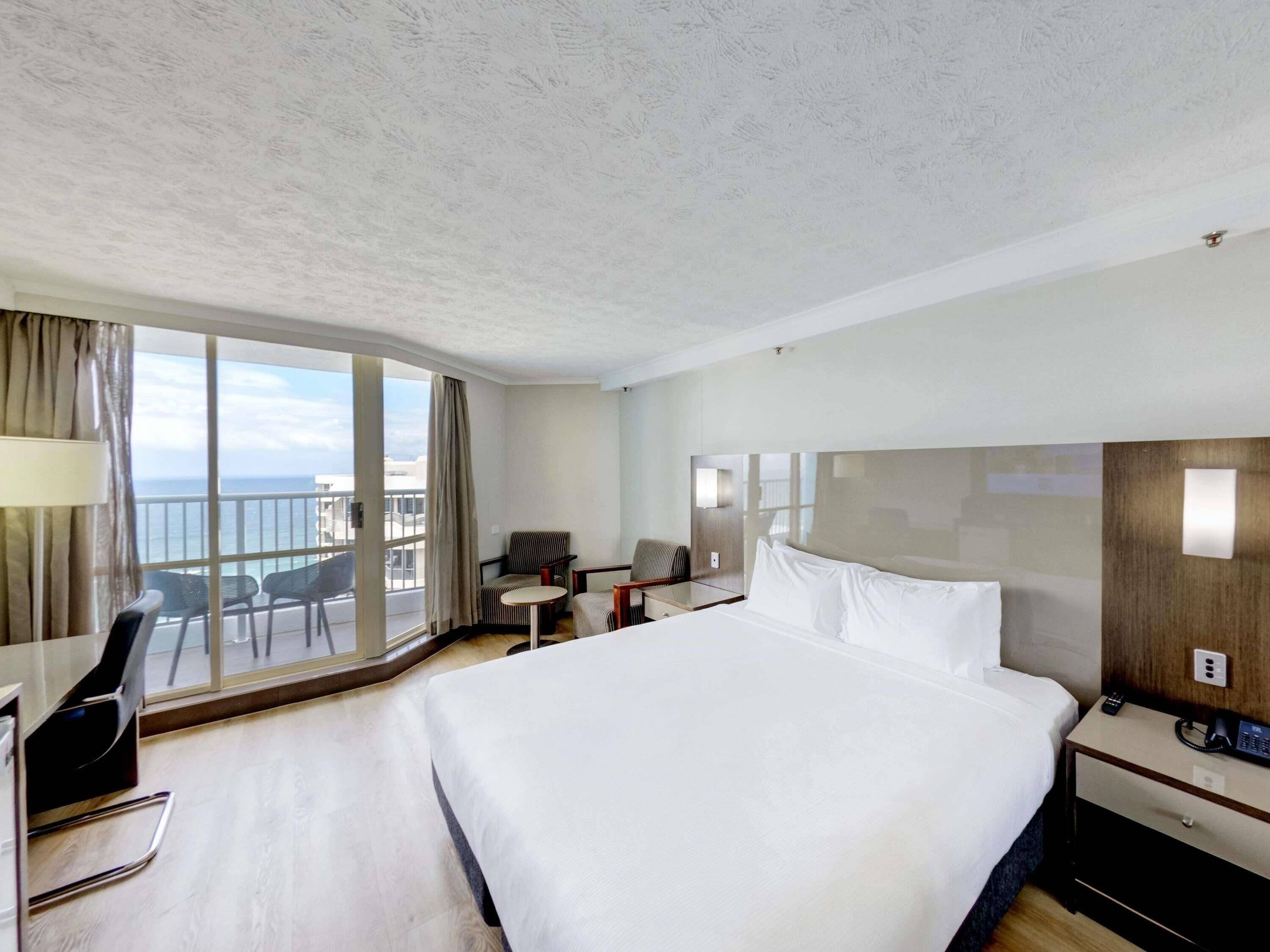 A hotel room with a large bed, a desk, a chair, a small table, and a balcony with a view of the sea.