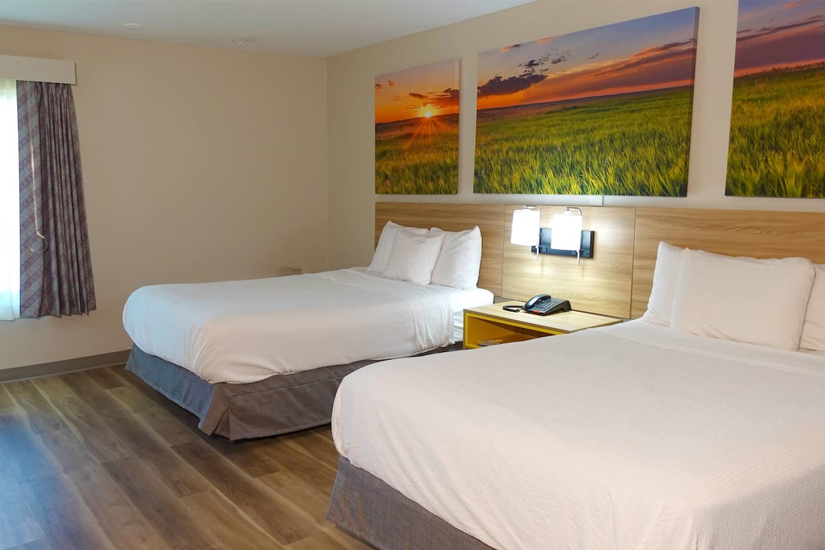 Une chambre d’hôtel avec deux lits, une tête de lit en bois, une table de chevet avec une lampe et deux tableaux représentant des paysages agricoles, accrochés au mur.