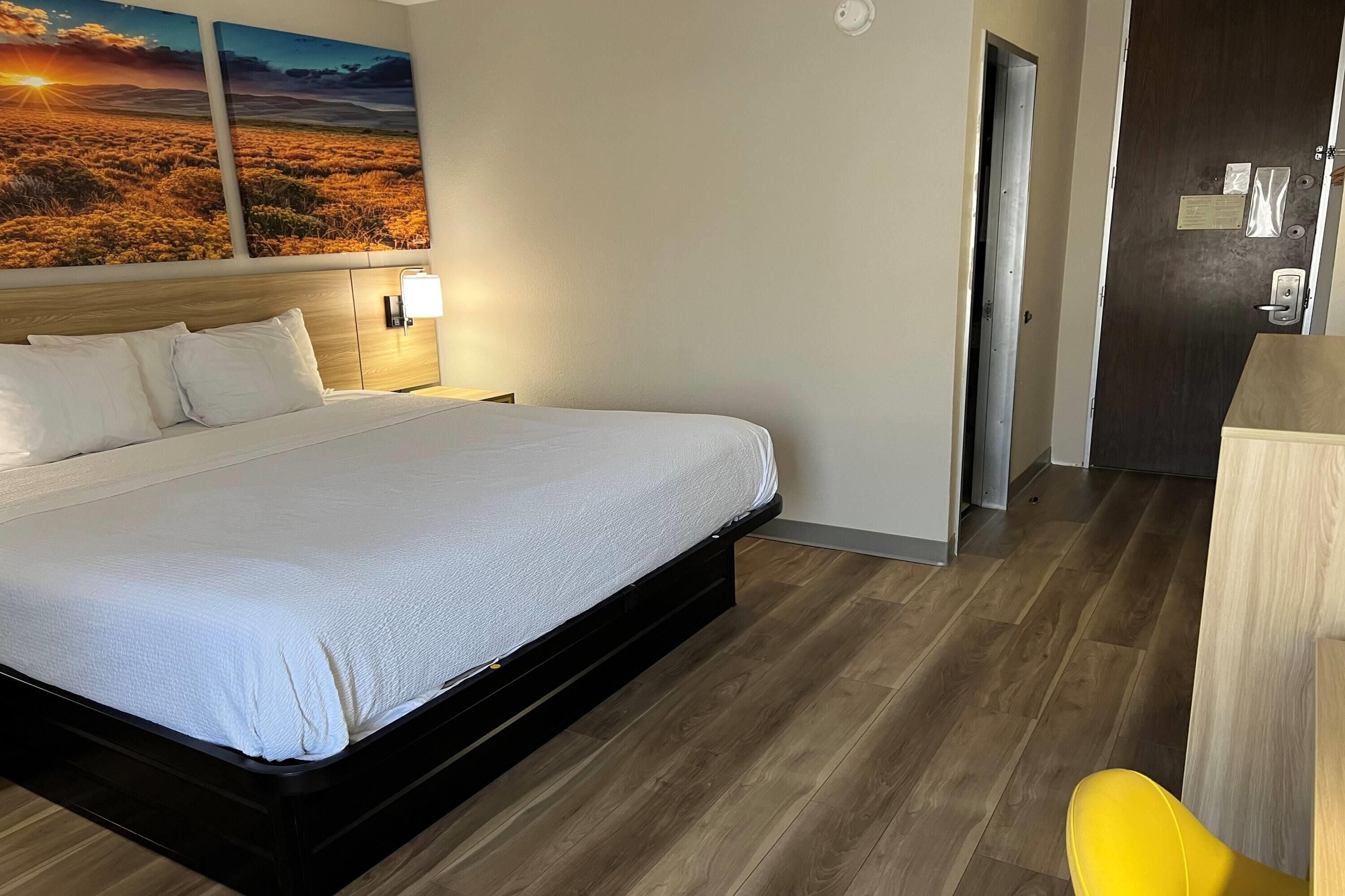 A hotel room with a bed, a nightstand with a lamp, a wooden floor, and a door.