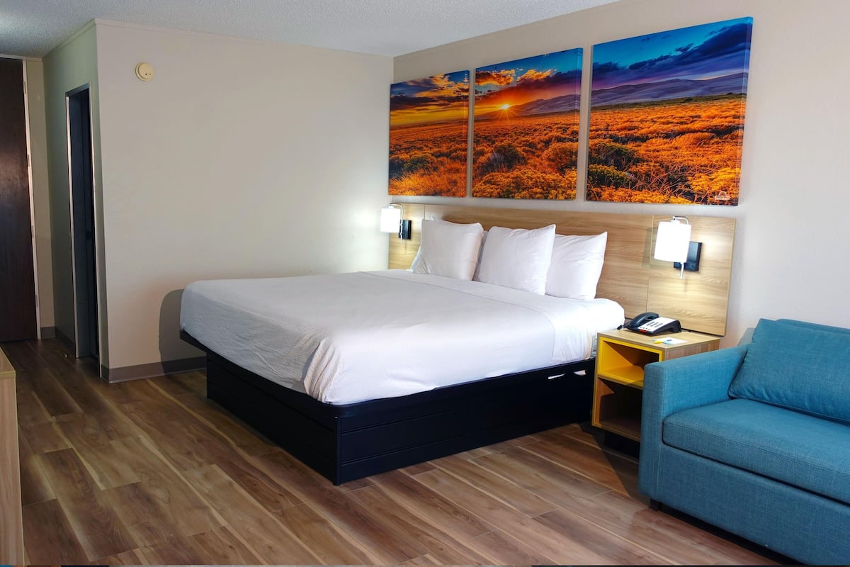 Une chambre d’hôtel avec un lit, une table de chevet avec un téléphone, un fauteuil bleu et un mur orné de trois tableaux représentant des couchers de soleil.