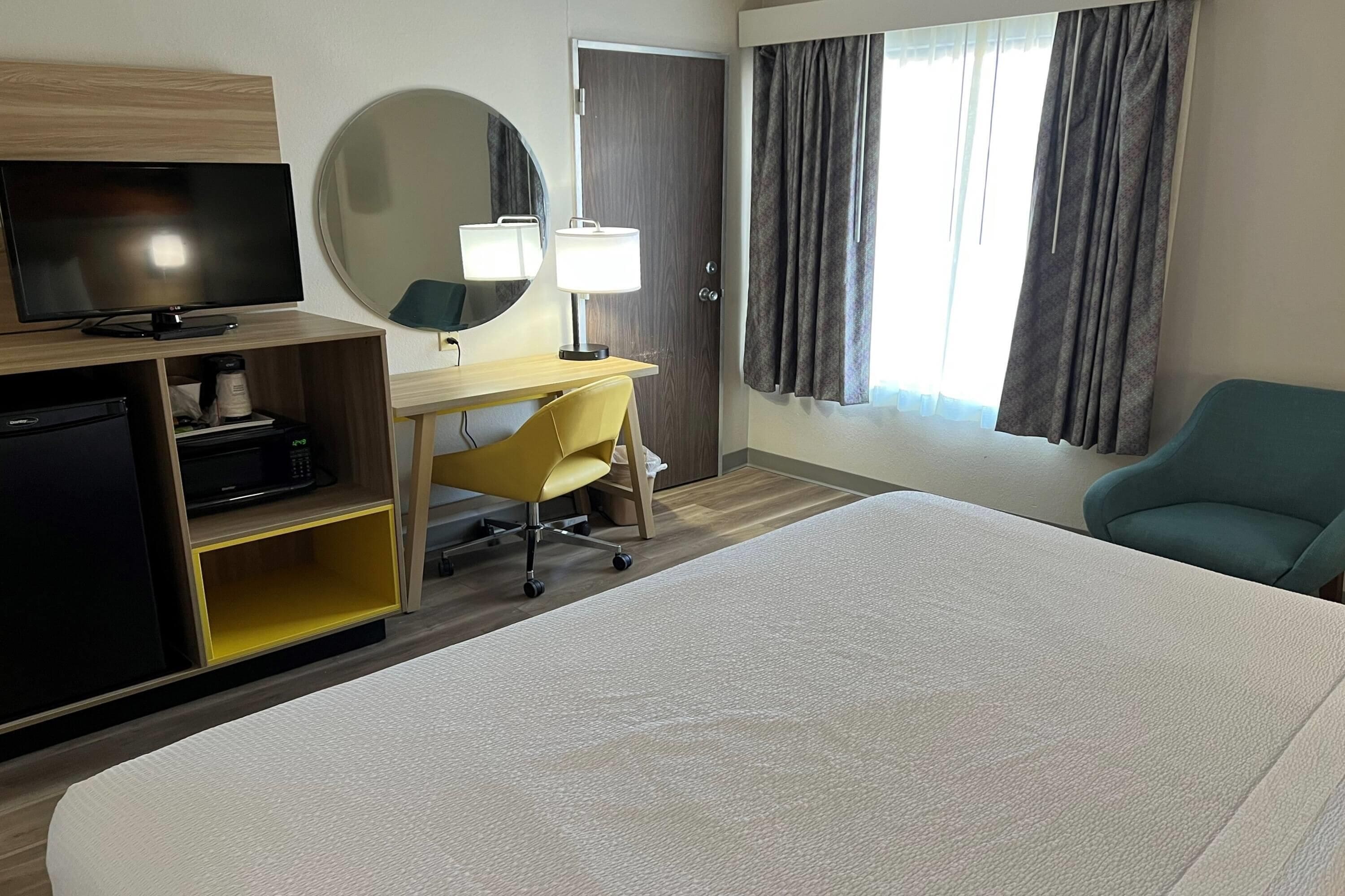 A hotel room with a bed, a desk with a chair, a television, a mirror, and a window with curtains.