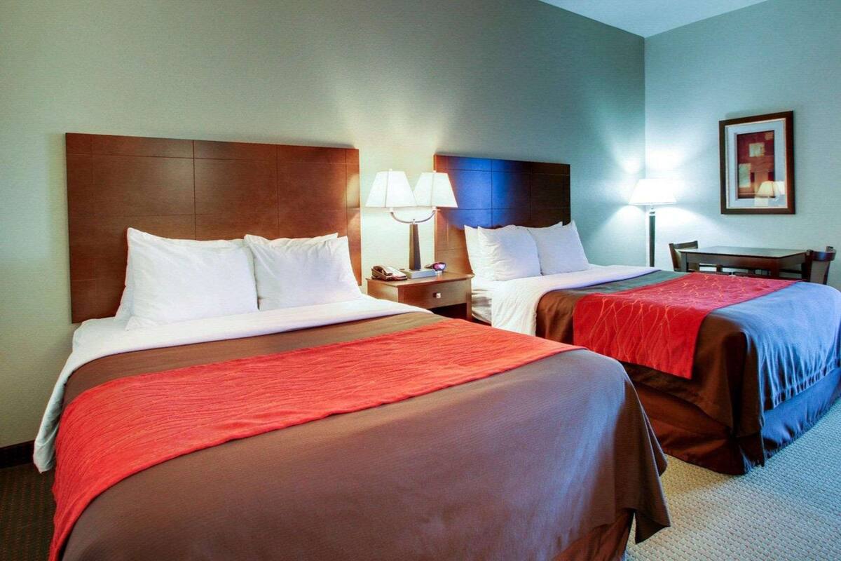 A hotel room with two beds, each with a wooden headboard, white bedding, and red throw blankets. There are bedside tables with lamps, a framed picture on the wall, and a small table with chairs.