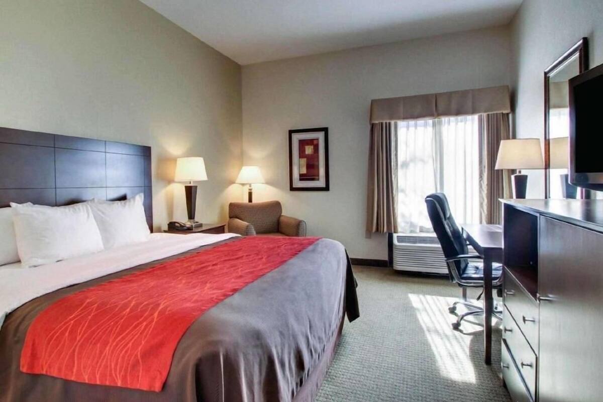 A hotel room with a bed, two bedside lamps, a desk with a chair, a television, and an air conditioning unit.