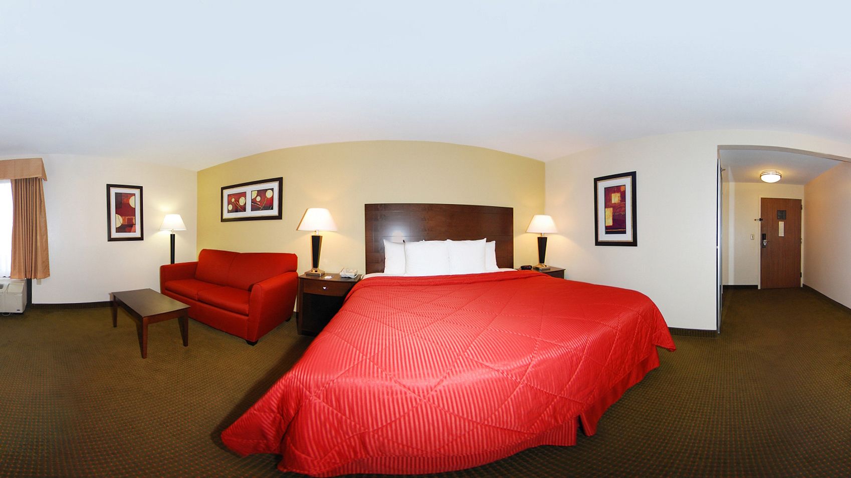 A hotel room with a large bed, a red sofa, a small table, two bedside lamps, and a door leading to another room.