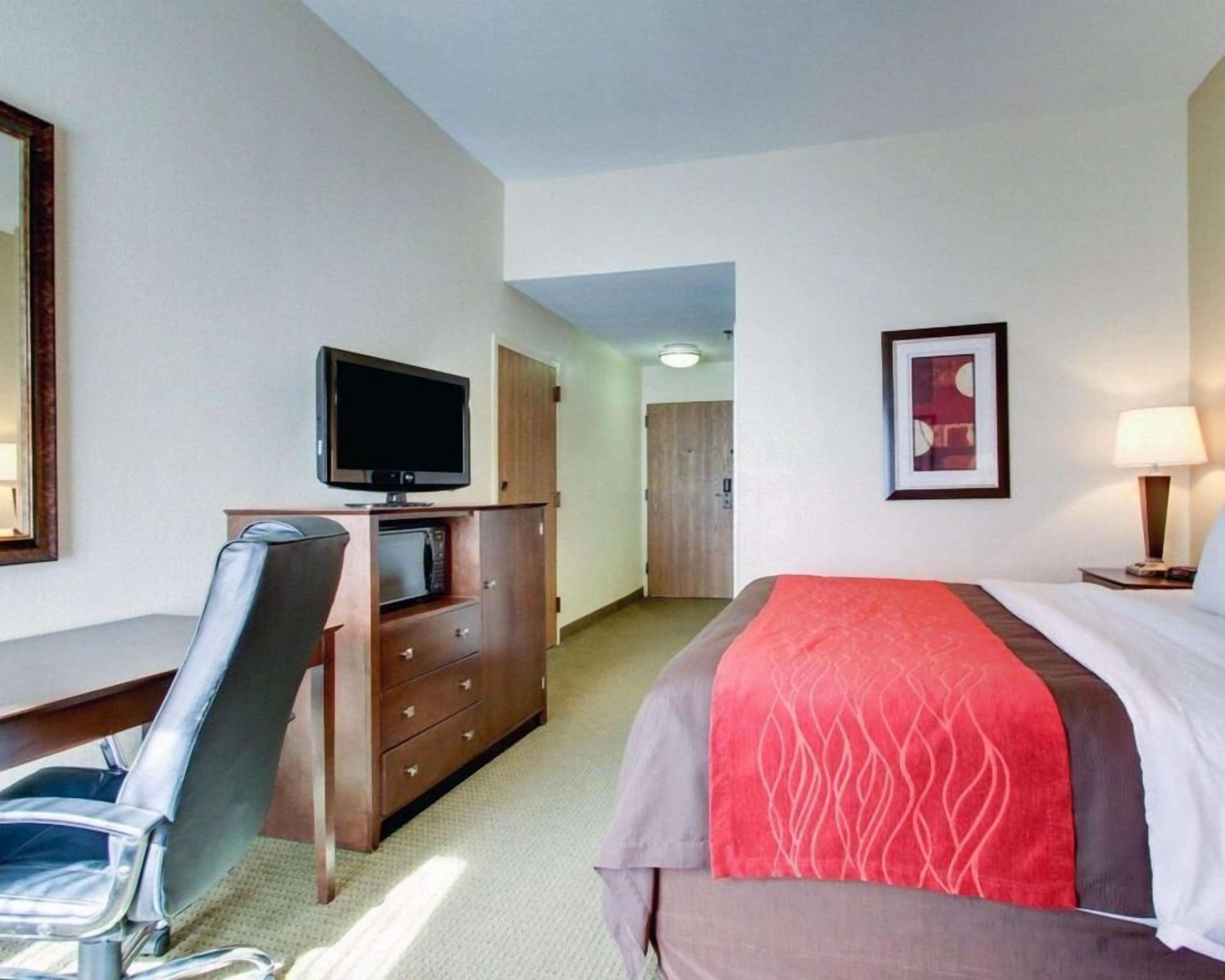 A hotel room with a bed, a desk with a chair, a television, a dresser, and a framed picture on the wall.