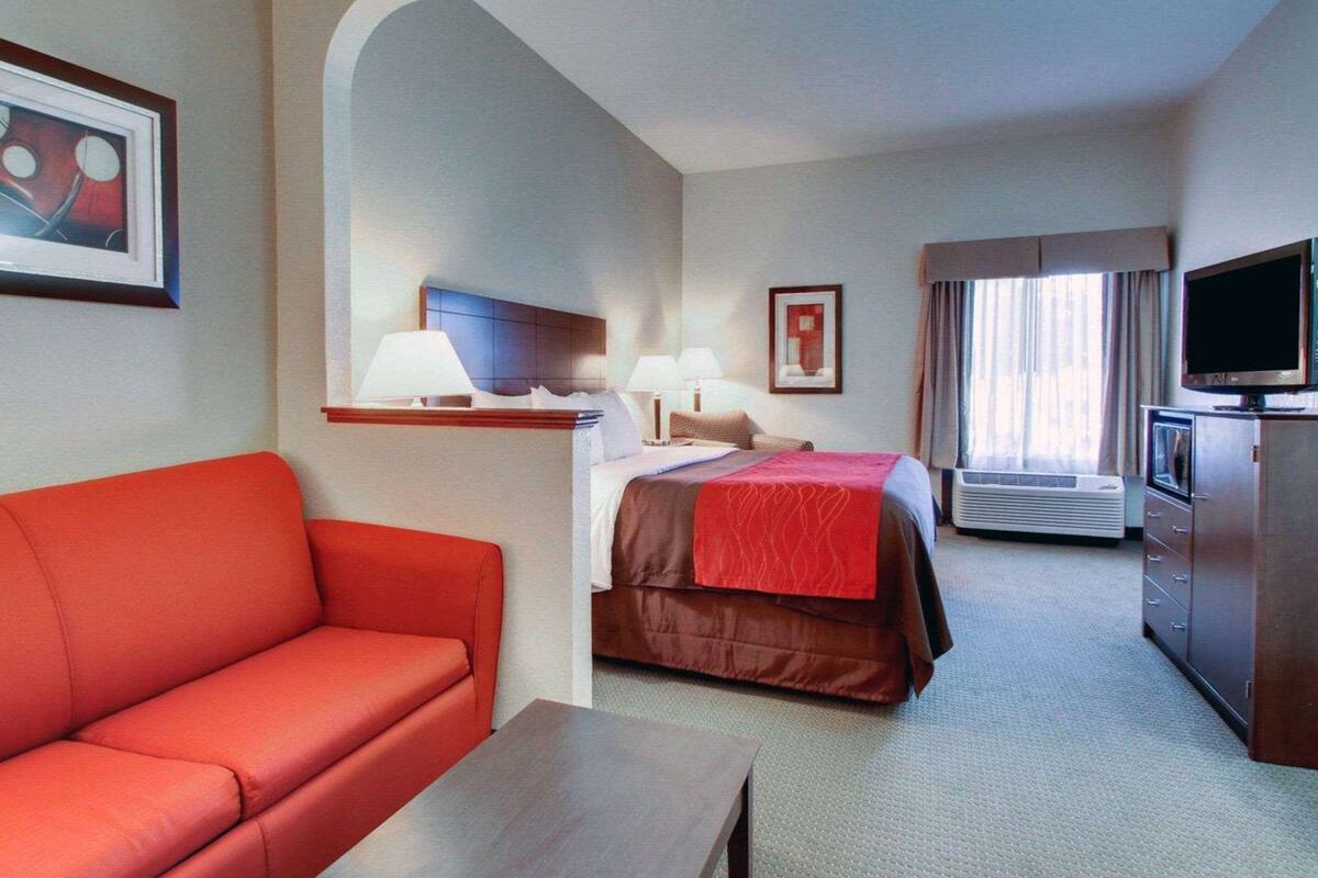 A hotel room with a red sofa, a bed with a red bedspread, a TV, a desk, and a window with curtains.