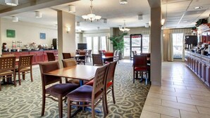 Ruime hotel eetruimte met meerdere tafels en stoelen, een keuken op de achtergrond, en grote ramen die natuurlijk licht binnenlaten.