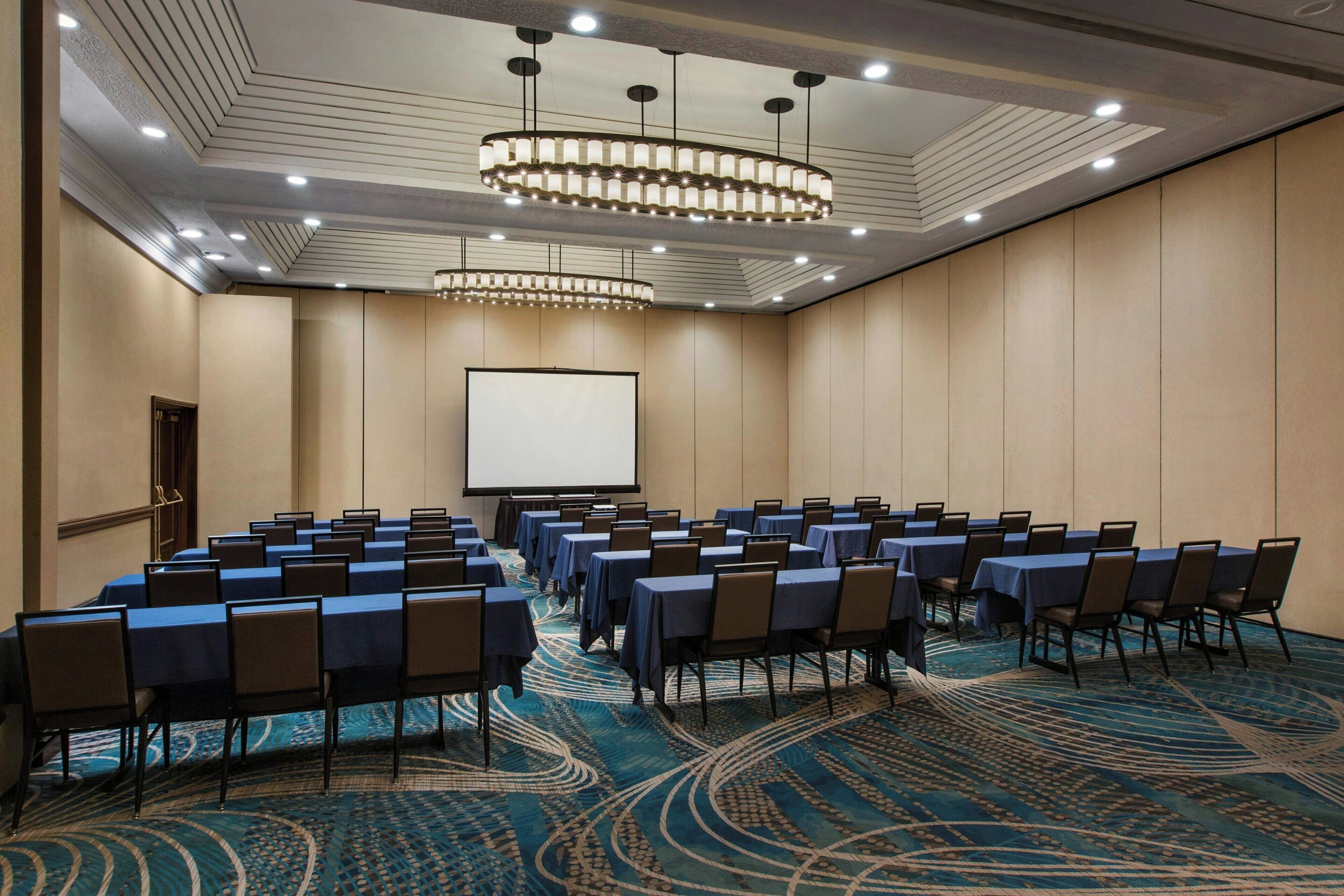 A conference room with rows of tables and chairs, a large screen, and a patterned carpet.