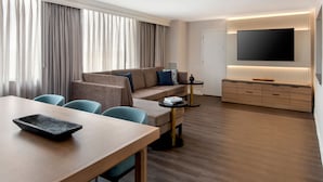 A modern hotel room with a wooden dining table, a sofa, a TV mounted on the wall, and a large window with curtains. - Buffalo Marriott Niagara (Buffalo)