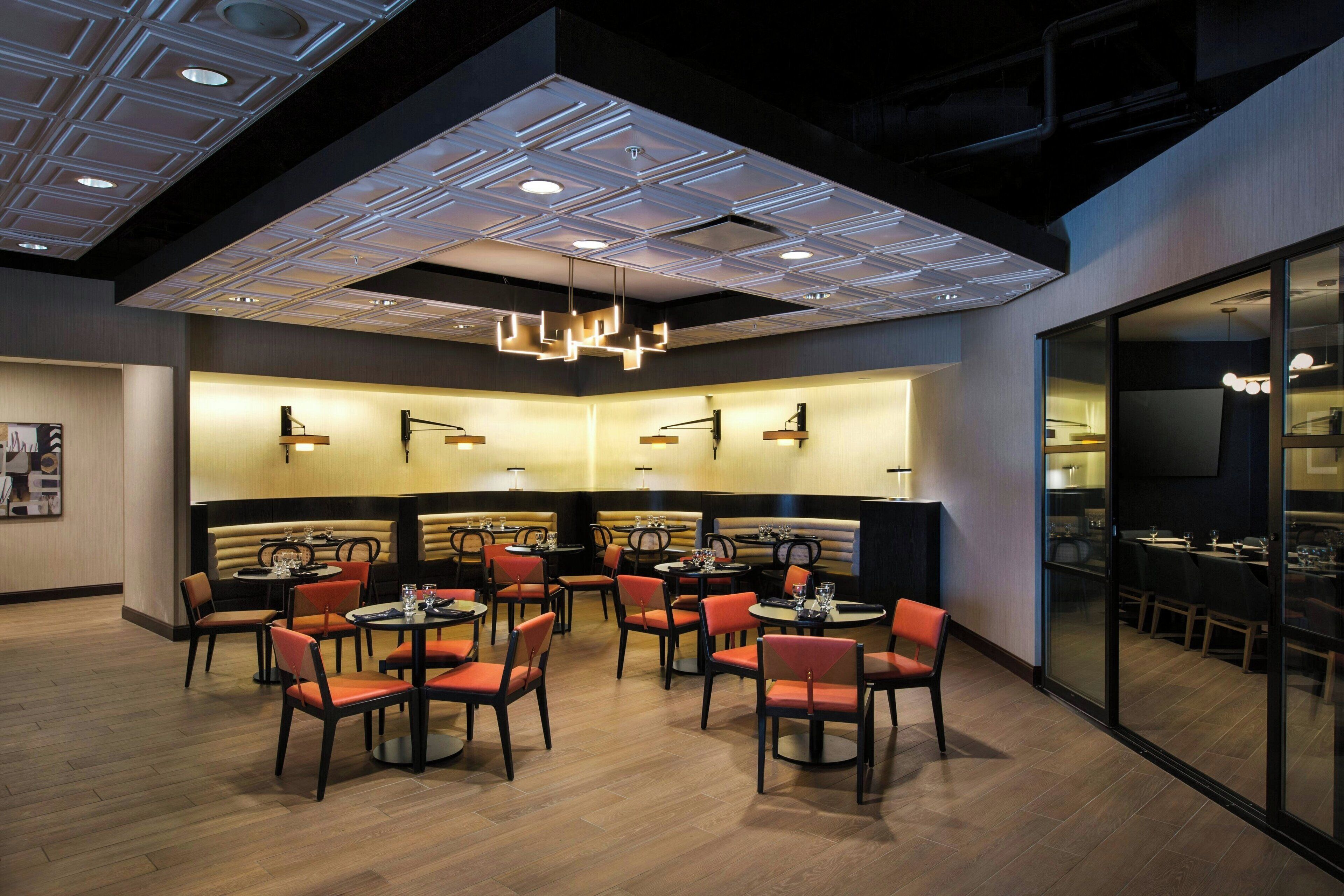 A modern restaurant with round tables and orange chairs, a high ceiling with recessed lighting, and a glass partition separating the dining area from the kitchen.