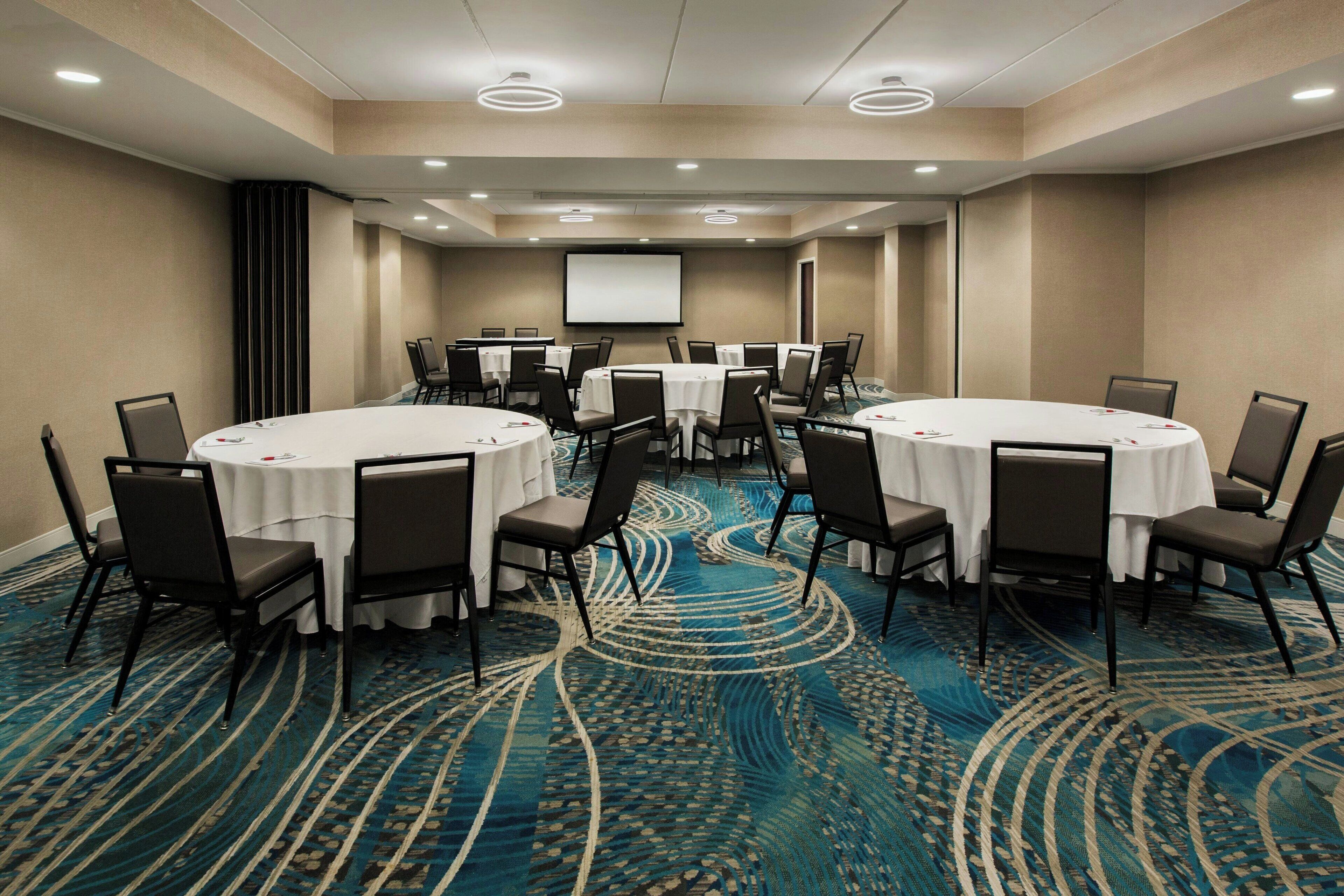 A conference room with round tables, chairs, a projector screen, and a carpeted floor.