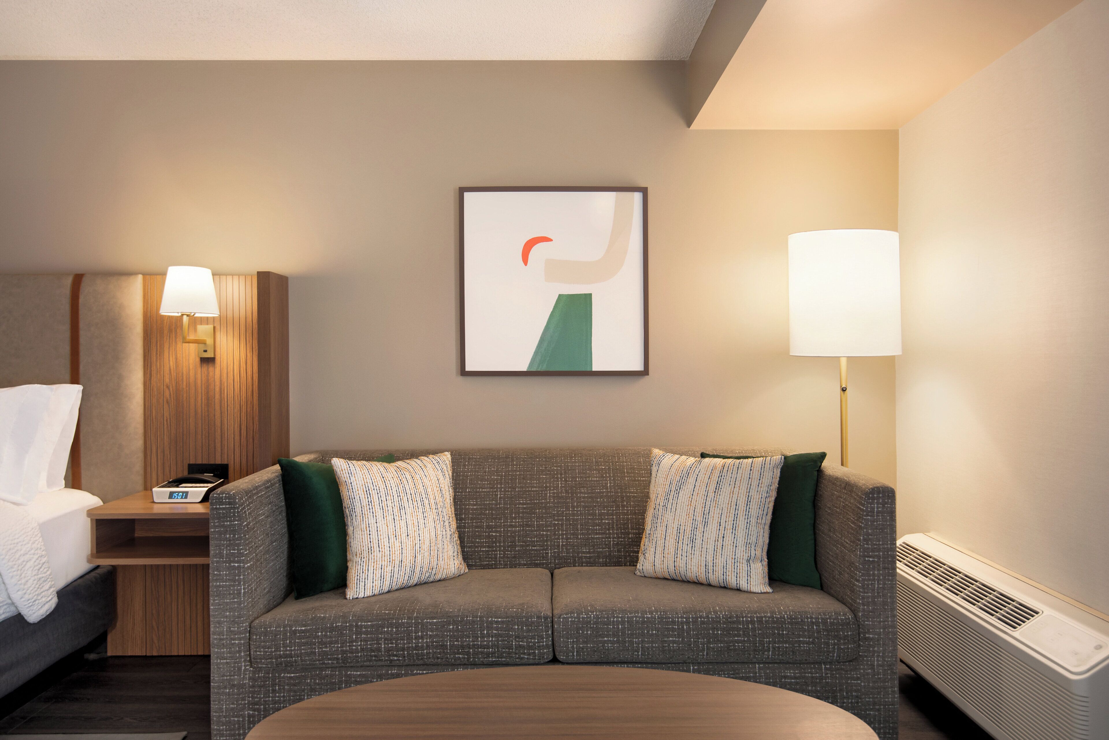 A hotel room with a grey sofa, two pillows, a small bedside table with a lamp, a framed abstract artwork, and a radiator.