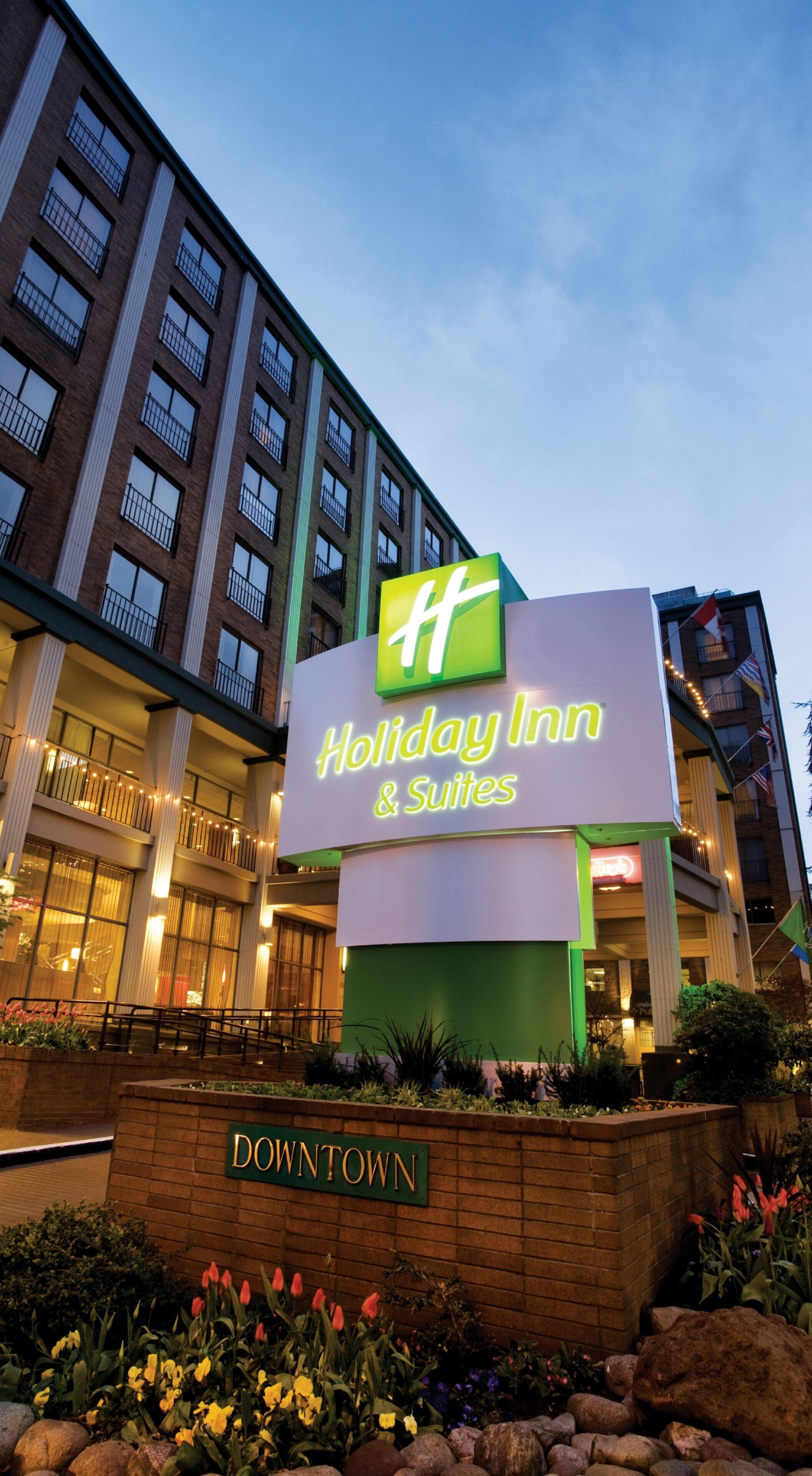 Ein Schild des Holiday Inn Hotels mit grün-weißem Logo, gelegen in einem Stadtgebiet mit einer Fassade aus Backsteinen und einem begrünten Eingangsbereich.