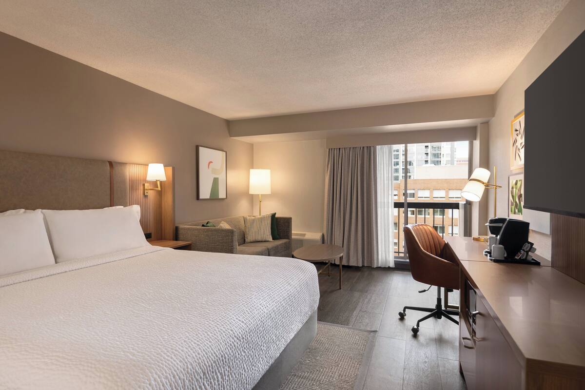 Quarto de hotel com uma cama grande, uma escrivaninha com computador, uma cadeira, um sofá e uma janela com cortinas.
