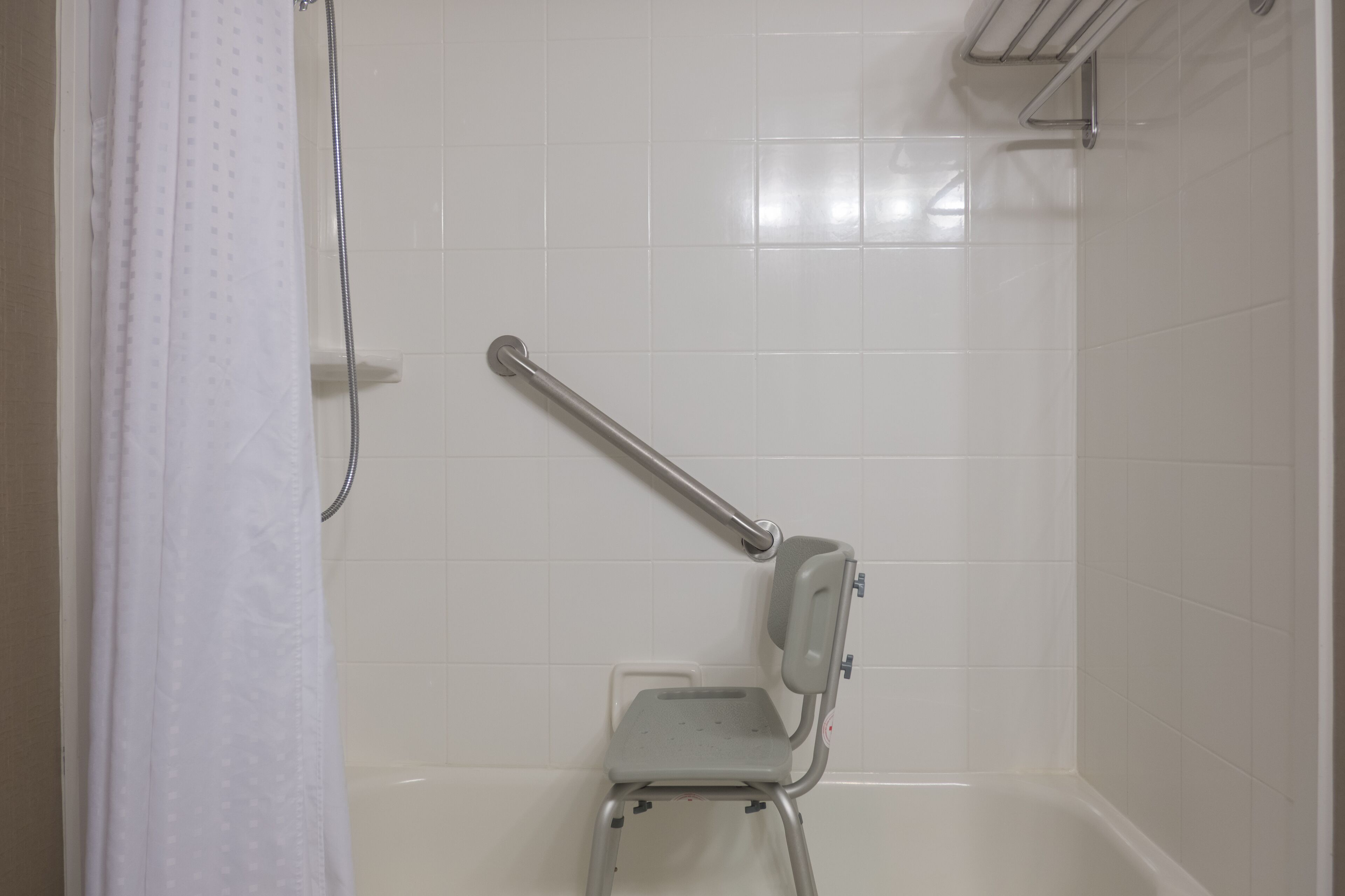 Zona de ducha con barra de apoyo, asiento de ducha y pared de azulejos blancos.
