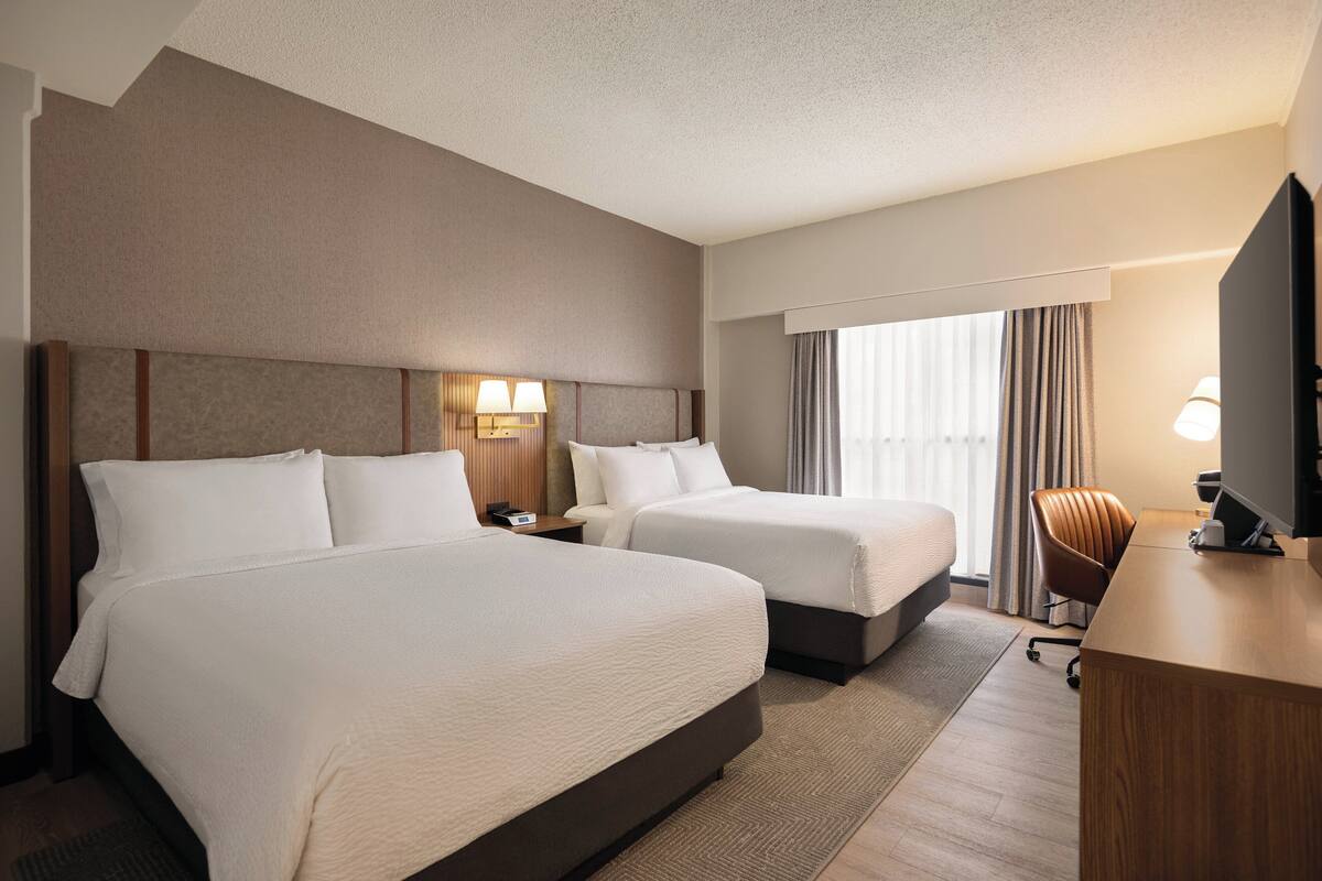 Quarto de hotel com duas camas, uma escrivaninha, uma TV e uma janela com cortinas.