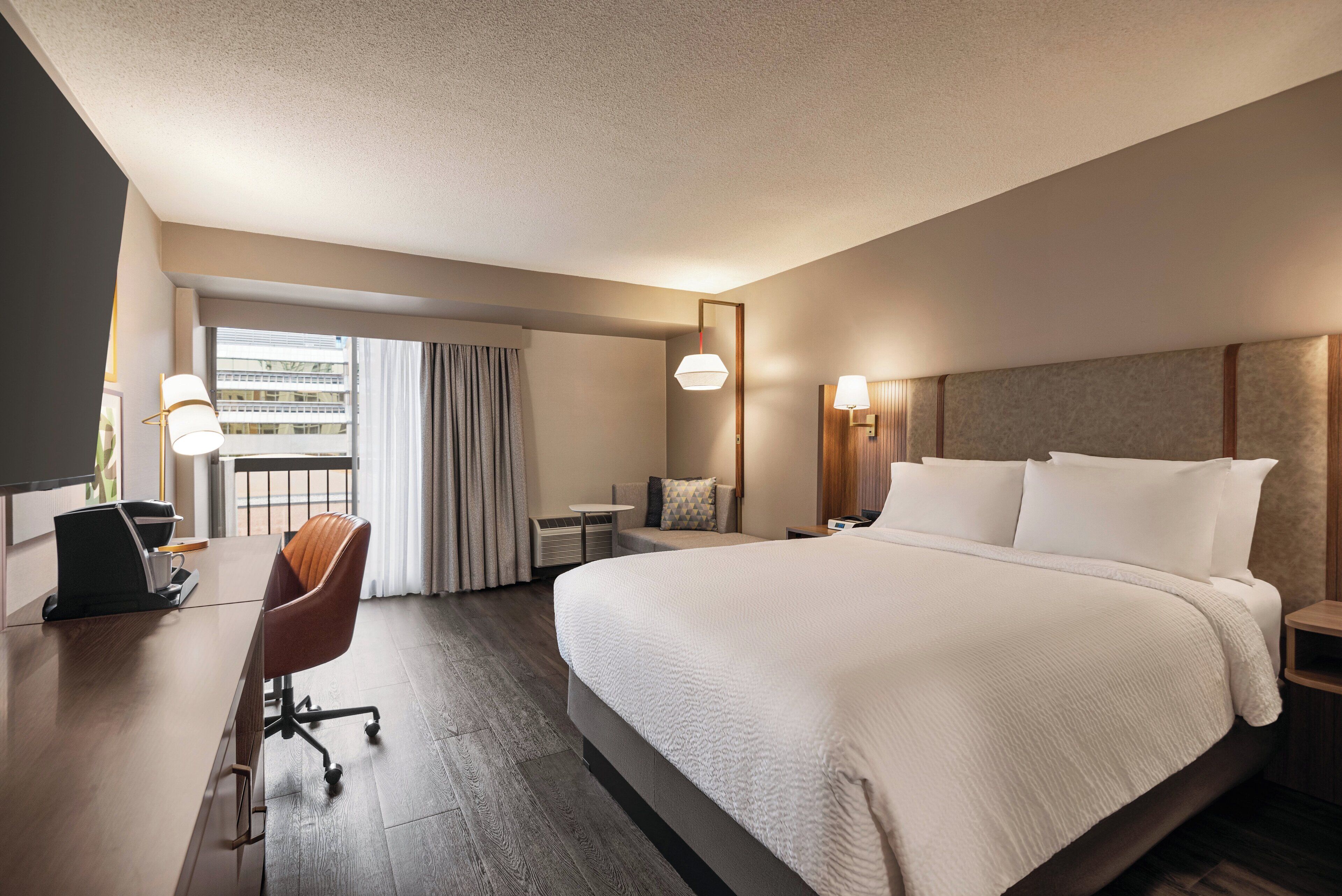 A hotel room with a large bed, a desk with a chair, a small table, and a window with curtains.