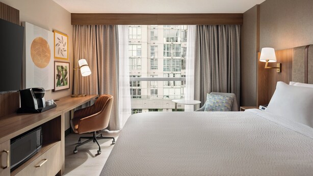 Quarto de hotel com uma cama grande, uma escrivaninha com cafeteira, uma cadeira, uma mesa pequena e vista para um edifício pela janela.