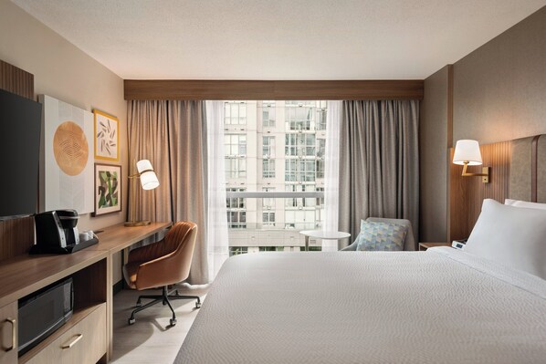 A hotel room with a large bed, a desk with a coffee maker, a chair, a small table, and a view of a building through the window. - Holiday Inn & Suites Vancouver Downtown by IHG (Vancouver)