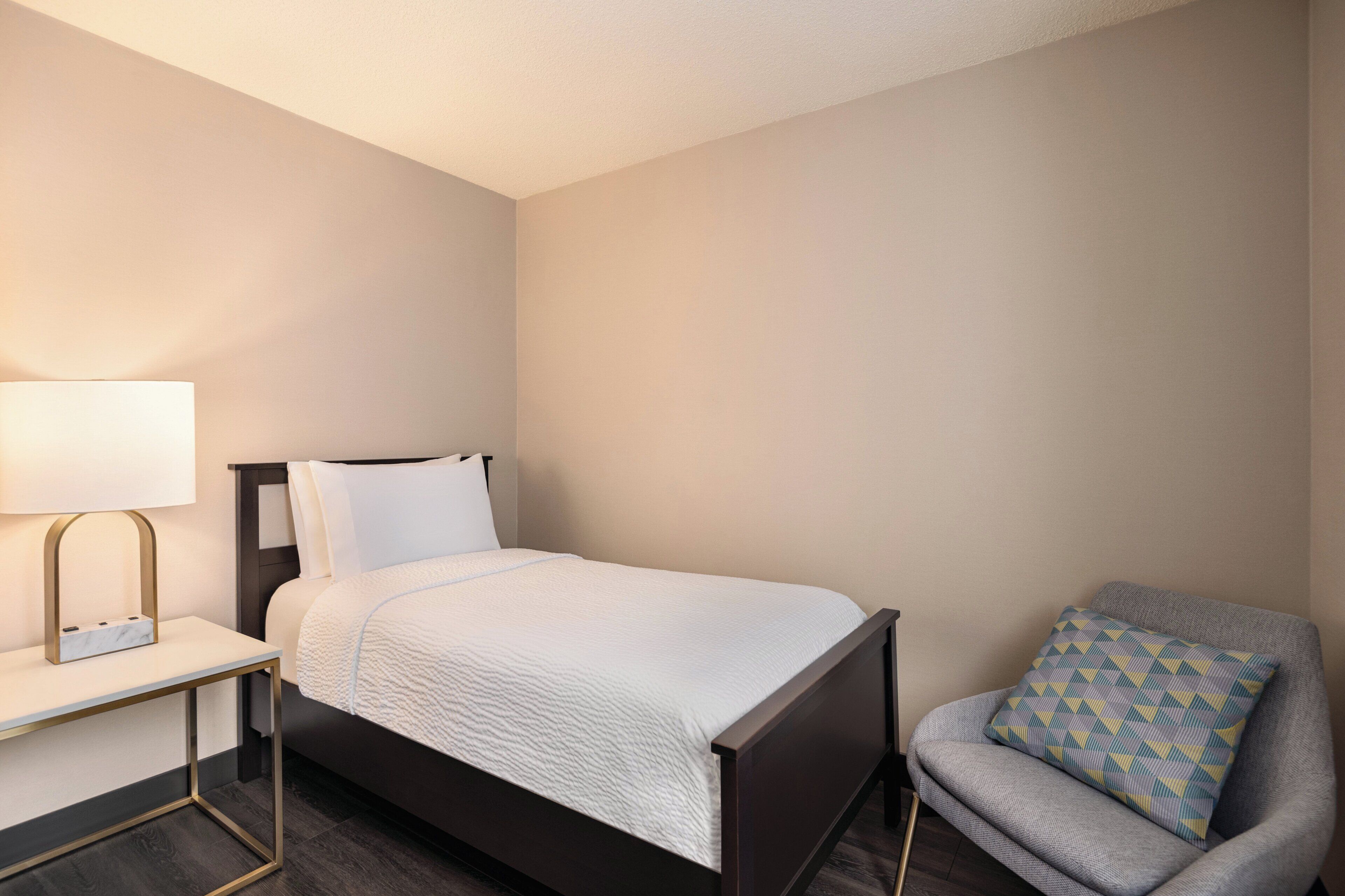 A neatly arranged hotel room with a bed, a chair, a bedside table, and a lamp.